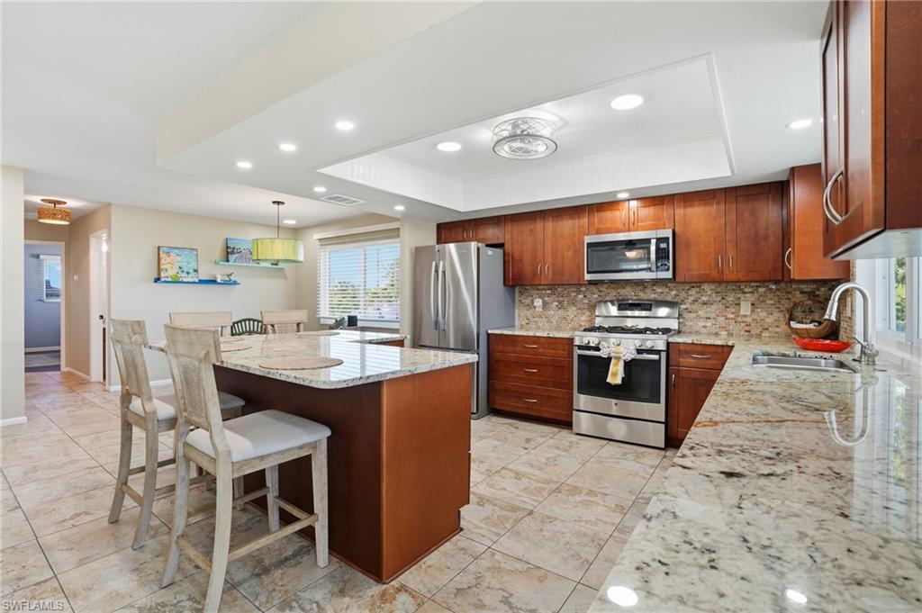 Kitchen with light stone countertops, stainless steel appliances, a kitchen bar, a raised ceiling, and recessed lighting - Virtually Edited Image