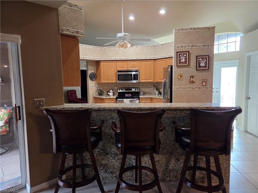 Kitchen featuring light tile patterned floors, appliances with stainless steel finishes, a kitchen bar, tile backsplash, and granite stone kick wall.