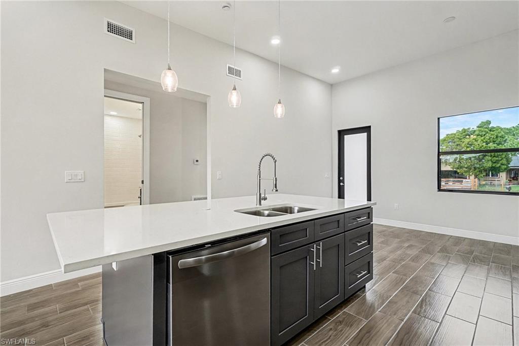 Kitchen with dishwasher, wood finish floors, hanging light fixtures, an island with sink, and light stone counters