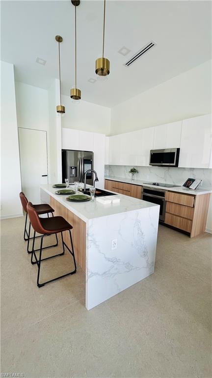 Kitchen featuring a waterfall island with a stone-finish countertop, sleek white upper cabinetry, light wood-finish lower cabinetry, and stainless steel appliances