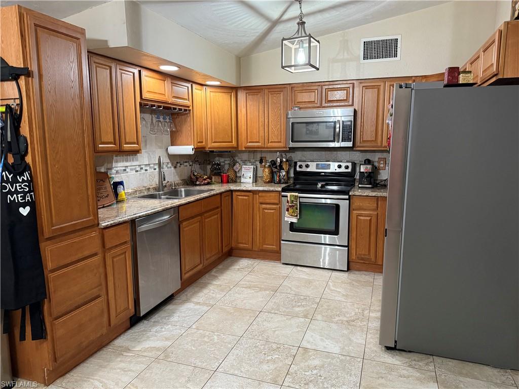 Kitchen featuring stainless steel appliances, brown cabinetry, vaulted ceiling, backsplash, and light stone counters