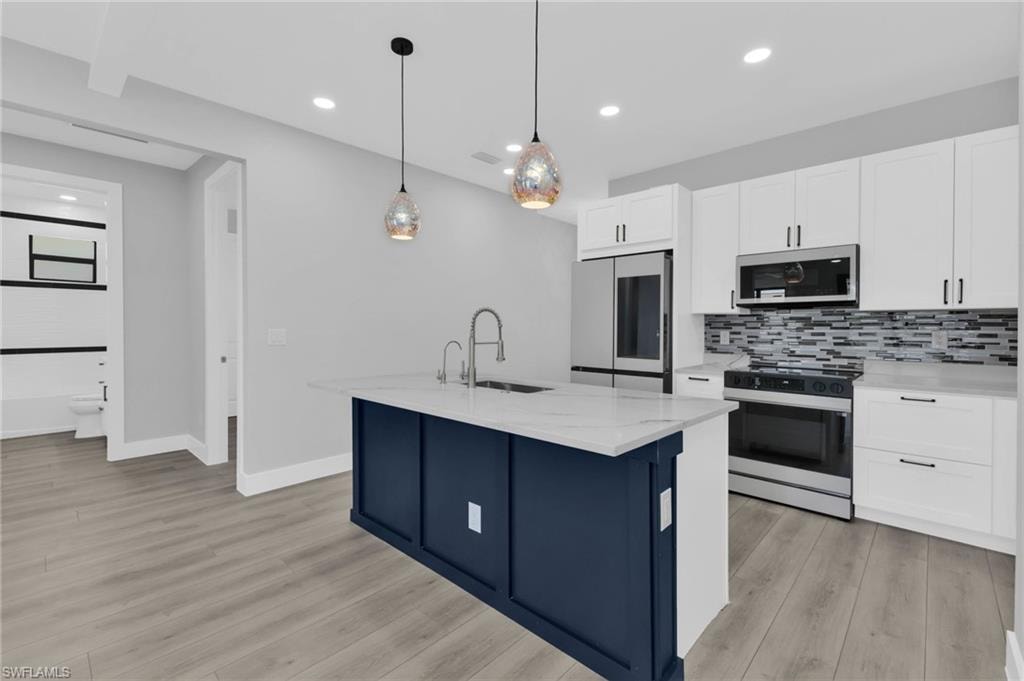 Kitchen with appliances with stainless steel finishes, white cabinets, light stone countertops, a center island with sink, and decorative light fixtures