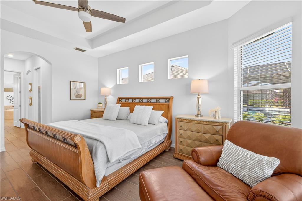 Bedroom with wood tiled floors, a ceiling fan, a tray ceiling, arched walkways, and connected bathroom