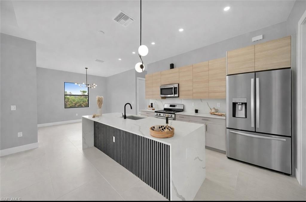 Kitchen featuring decorative light fixtures, stainless steel appliances, light brown cabinetry, light stone countertops, and a kitchen island with sink