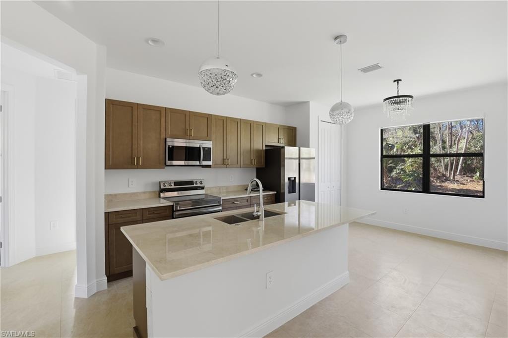 Kitchen featuring appliances with stainless steel finishes, hanging light fixtures, a center island with sink, light stone countertops, and brown cabinetry