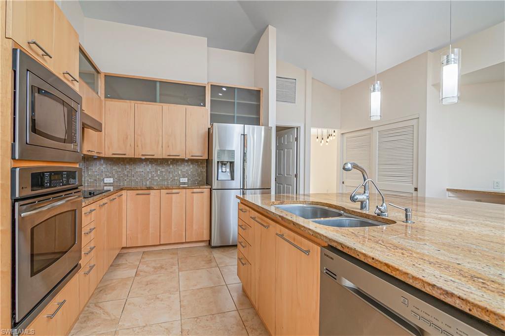Kitchen with glass insert cabinets, light stone counters, light brown cabinets, appliances with stainless steel finishes, and hanging light fixtures