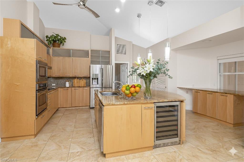 Kitchen with light stone counters, hanging light fixtures, light brown cabinets, beverage cooler, and stainless steel appliances