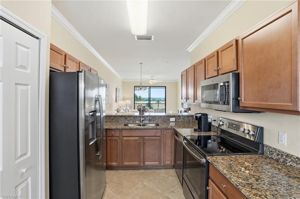 Kitchen featuring stainless steel appliances, crown molding, dark stone countertops, brown cabinetry, and decorative light fixtures