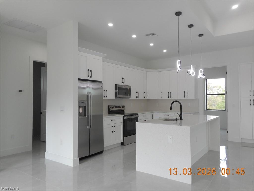 Kitchen featuring stainless steel appliances, white cabinets, a kitchen island with sink, decorative light fixtures, and light stone counters