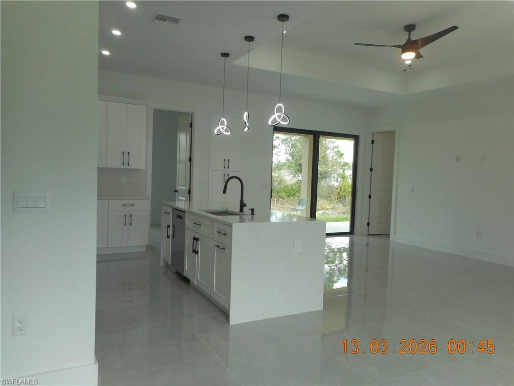 Kitchen featuring white cabinetry, an island with sink, light stone counters, hanging light fixtures, and a ceiling fan