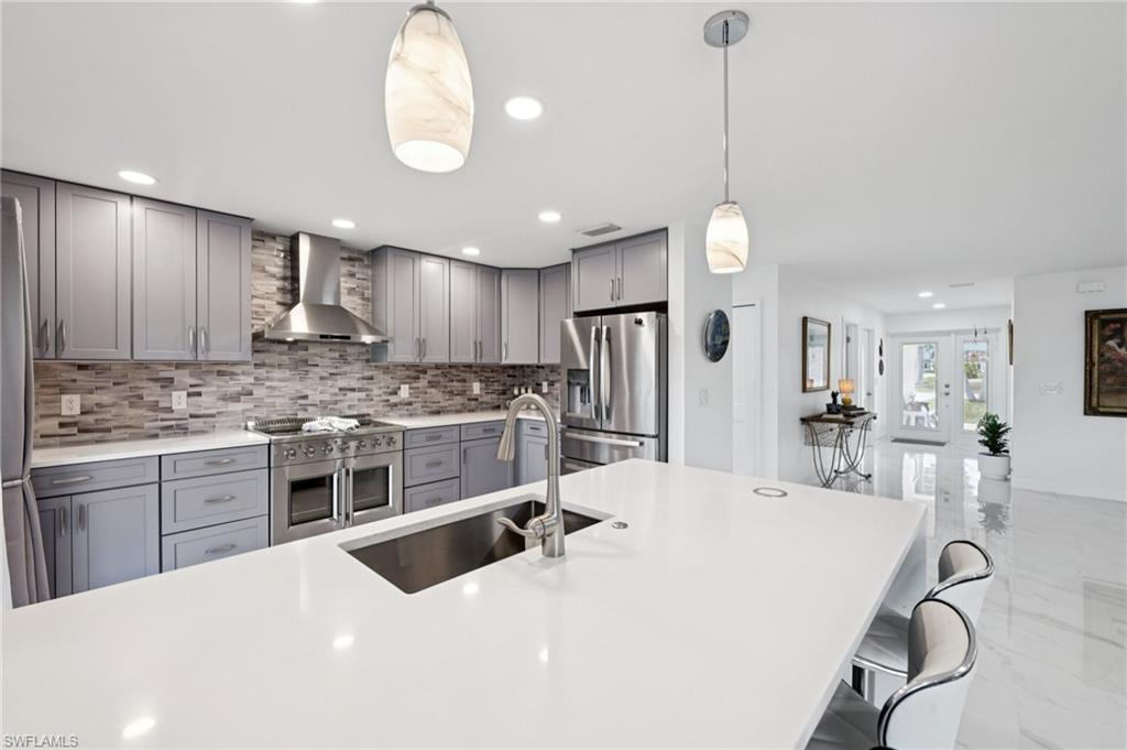 Kitchen featuring gray cabinetry, wall chimney range hood, stainless steel appliances, pendant lighting, and light stone counters