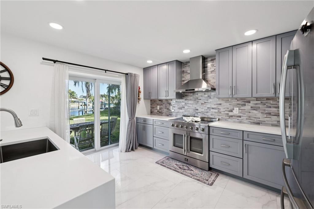 Kitchen with stainless steel appliances, gray cabinetry, wall chimney range hood, light marble finish floors, and light stone counters