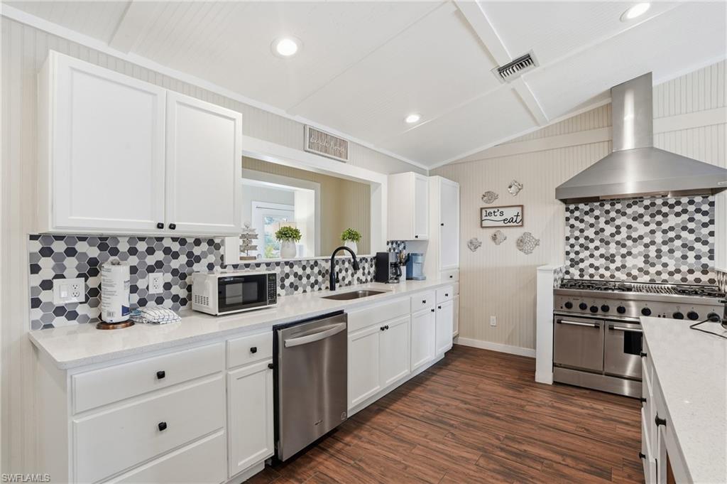 Kitchen featuring tasteful backsplash, white cabinetry, wall chimney range hood, appliances with stainless steel finishes, and lofted ceiling - Virtually Edited Image