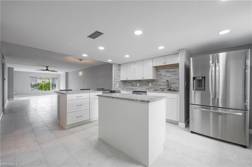 Kitchen featuring white cabinetry, stainless steel refrigerator with ice dispenser, a center island, hanging light fixtures, and light stone counters