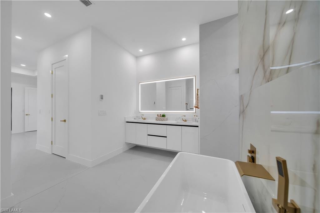 Freestanding soaking tub with brushed gold fixtures, a floating dual vanity with an illuminated mirror, large format wall tiles with subtle veining, recessed lighting, and light-toned flooring