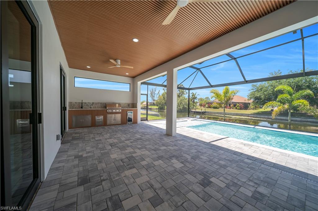 Expansive screened lanai featuring a built-in outdoor kitchen with stainless steel appliances, a wood-finish slatted ceiling with integrated lighting and ceiling fans, and a paver stone patio