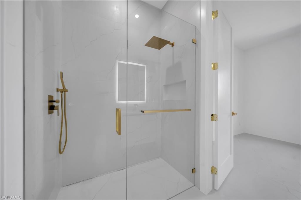 Contemporary glass-enclosed shower featuring large format white tile with subtle veining, a rainfall showerhead, handheld sprayer, and built-in shelving, all with gold-tone fixtures