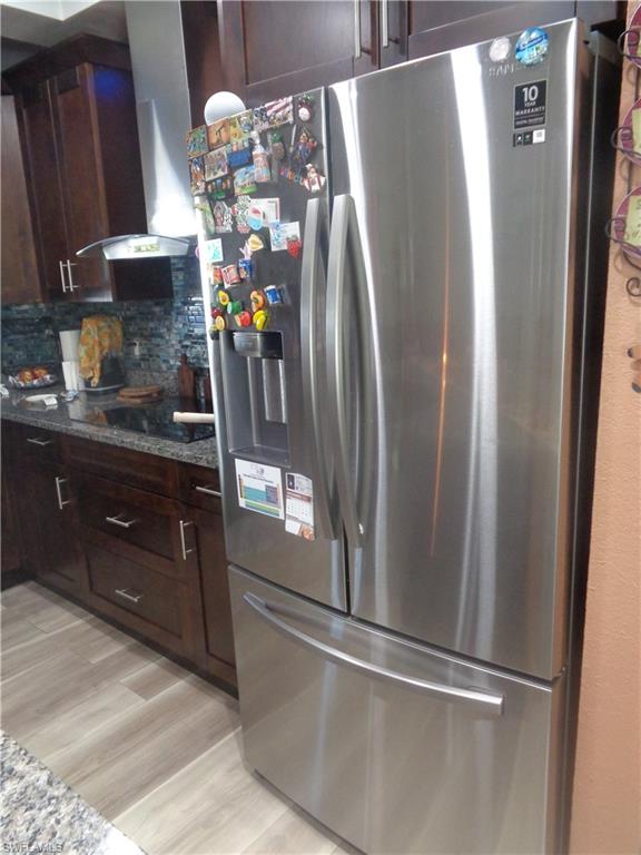 Kitchen with stainless steel side by side doors refrigerator with ice dispenser, dark brown cabinetry, granite countertops, wall chimney range hood, and backsplash