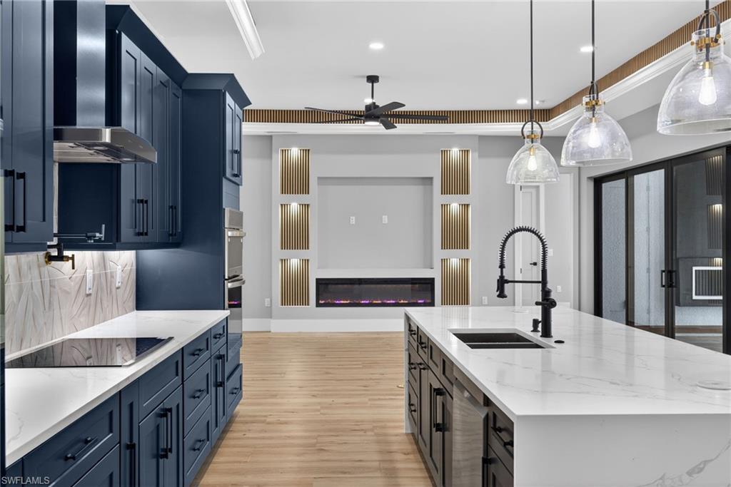 Kitchen featuring light stone countertops, wall chimney exhaust hood, hanging light fixtures, an island with sink, and ceiling fan