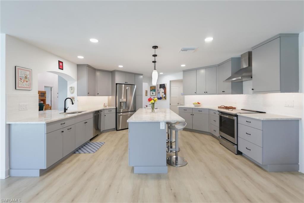 Kitchen featuring gray cabinetry, a kitchen breakfast bar, appliances with stainless steel finishes, wall chimney exhaust hood, and hanging light fixtures