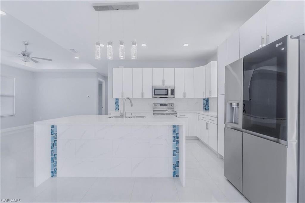 Kitchen featuring stainless steel appliances, a kitchen island with sink, white cabinetry, decorative light fixtures, and light stone counters