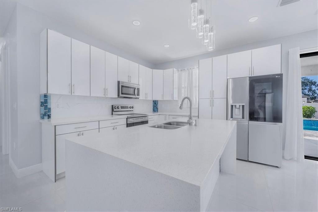Kitchen featuring stainless steel appliances, light stone counters, a kitchen island with sink, white cabinetry, and hanging light fixtures