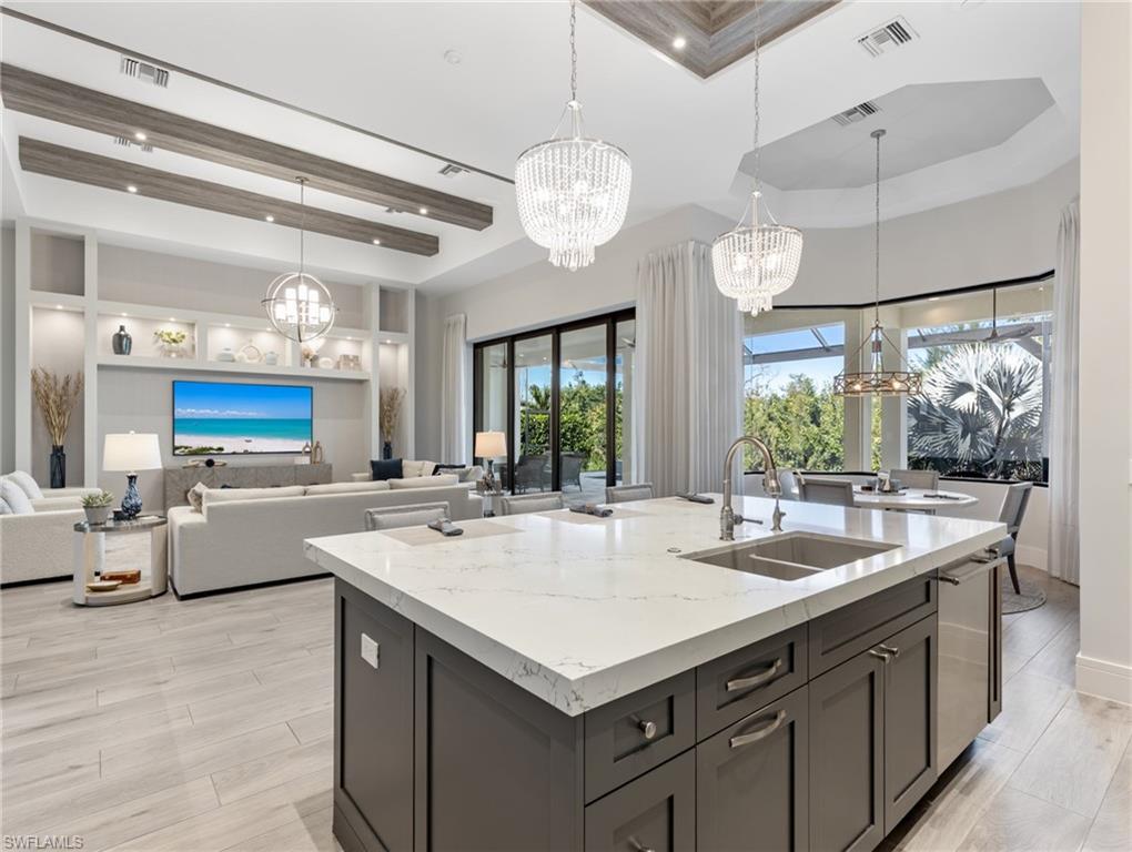 Kitchen featuring hanging light fixtures, a raised ceiling, light stone counters, a center island with sink, and a high ceiling