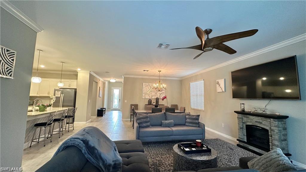 Living area with crown molding, light tile patterned floors, visible vents, a stone fireplace, and ceiling fan with notable chandelier