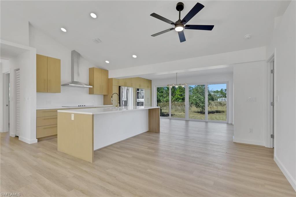 Kitchen featuring modern cabinets, wall chimney range hood, a center island with sink, light brown cabinetry, and light wood finished floors