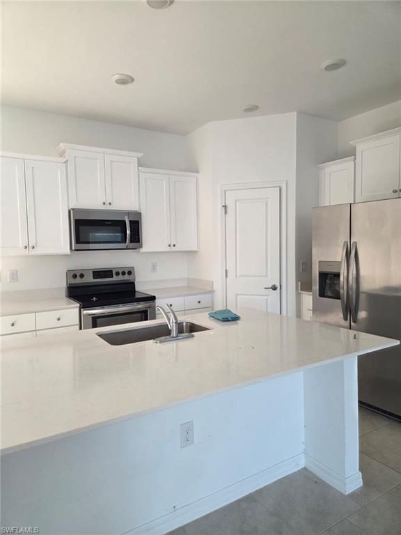 Kitchen with appliances with stainless steel finishes, white cabinets, light stone counters, and an island with sink