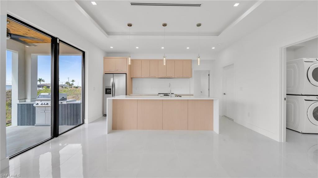 Kitchen featuring light brown cabinetry, pendant lighting, a center island with sink, stainless steel refrigerator with ice dispenser, and stacked washing machine and dryer