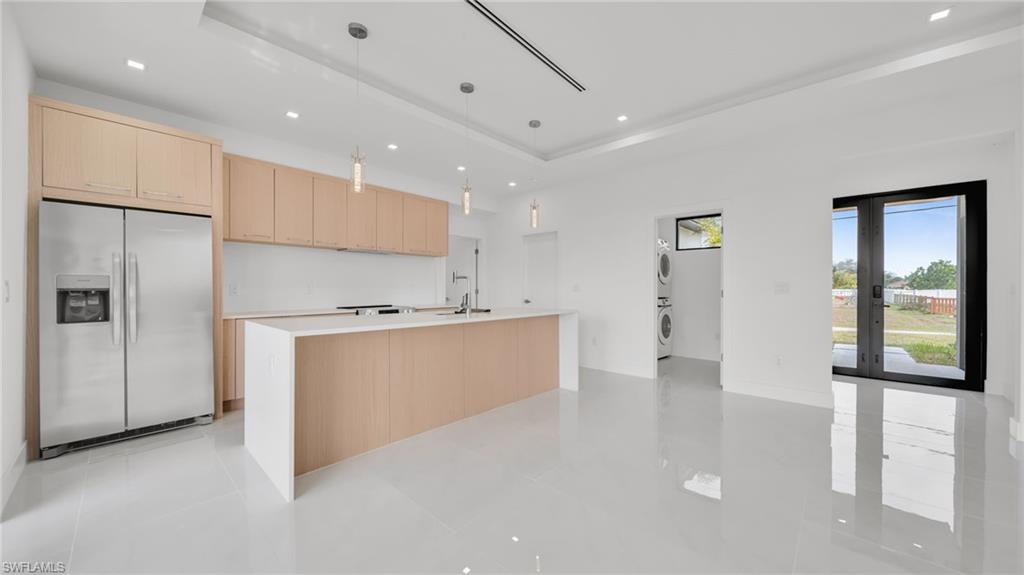 Kitchen with light brown cabinets, stainless steel fridge, decorative light fixtures, estacked washer and dryer, and an island with sink