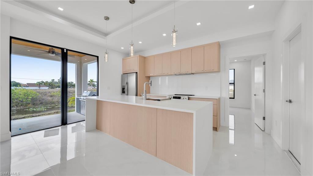 Kitchen with light brown cabinets, pendant lighting, light stone countertops, an island with sink, and stainless steel fridge