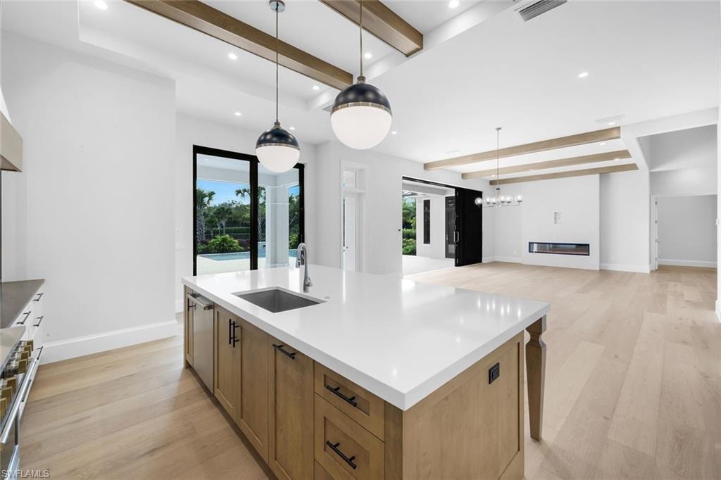 Kitchen with beam ceiling, light wood-type flooring, a kitchen island with sink, stainless steel range, and light stone counters