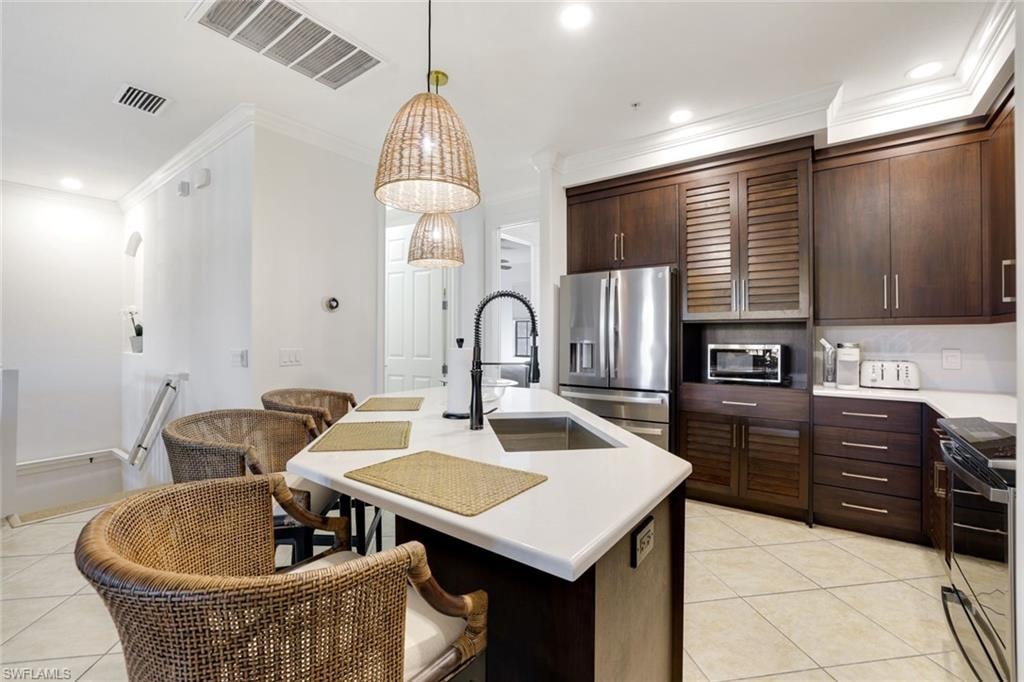Kitchen featuring dark wood finish cabinets, stainless steel appliances, crown molding, light tile patterned floors, and an island with sink
