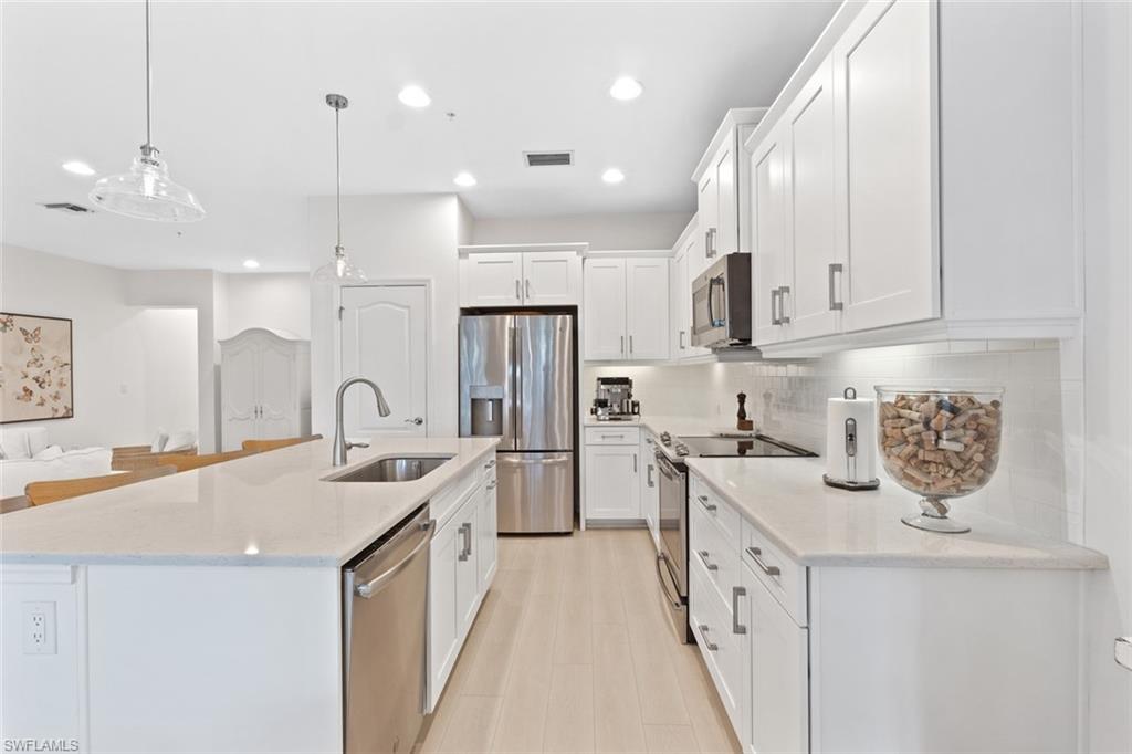 Kitchen featuring stainless steel appliances, hanging light fixtures, white cabinetry, light stone counters, and an island with sink