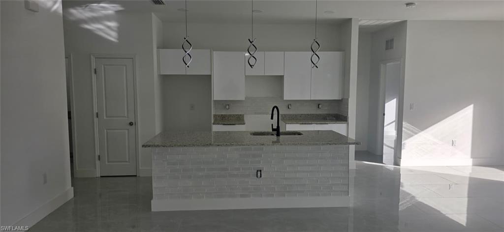 Kitchen with light stone countertops, white cabinetry, an island with sink, pendant lighting, and backsplash