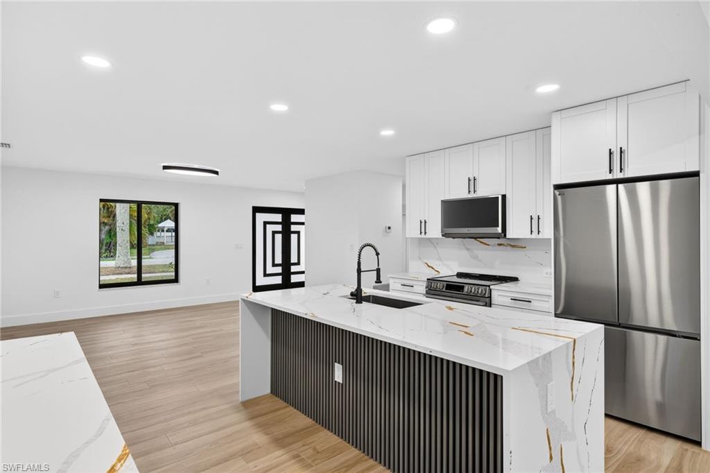 Kitchen with stainless steel appliances, light stone counters, white cabinets, an island with sink, and recessed lighting