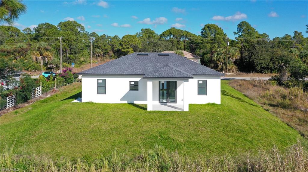 Back of house with stucco siding, a patio, a shingled roof, a yard, and view of wooded area