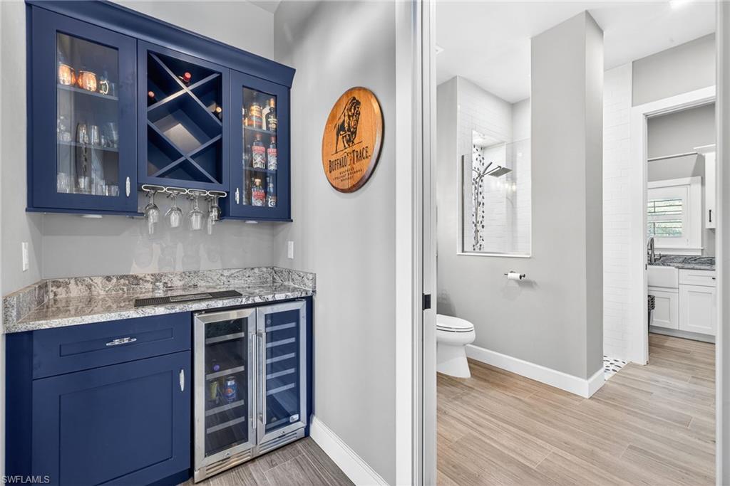 Indoor dry bar featuring blue cabinets, light stone counters, glass insert cabinets, beverage cooler, and light wood-style floors