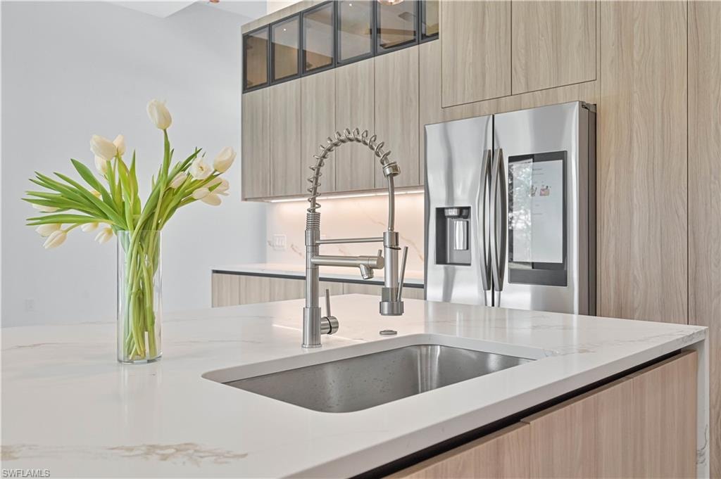 Kitchen featuring modern cabinets, stainless steel fridge with ice dispenser, light wood finish cabinets, light stone counters, and glass fronted cabinets