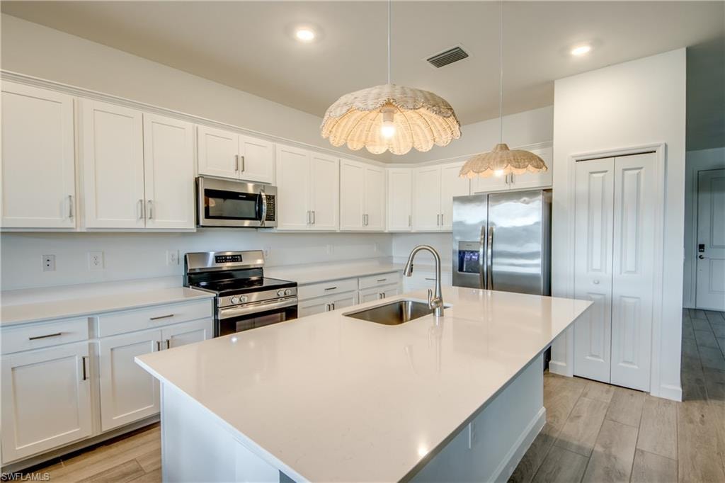 Kitchen featuring appliances with stainless steel finishes, white cabinets, light wood-style floors, a center island with sink, and hanging light fixtures