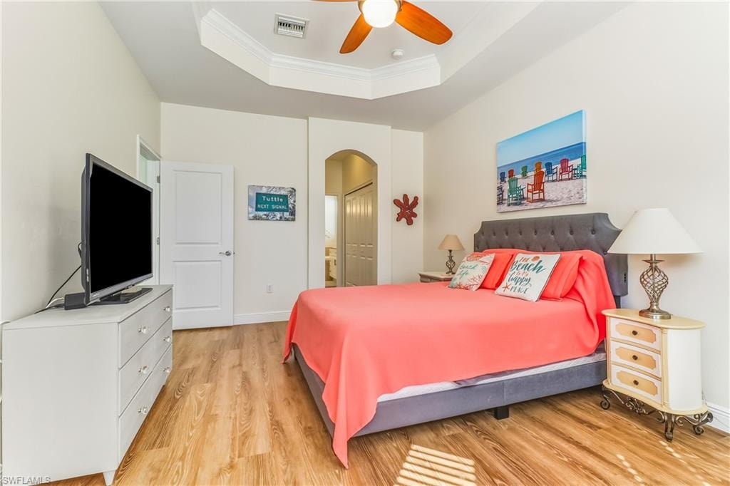 Bedroom featuring ornamental molding, light wood-style flooring, arched walkways, connected bathroom, and a tray ceiling