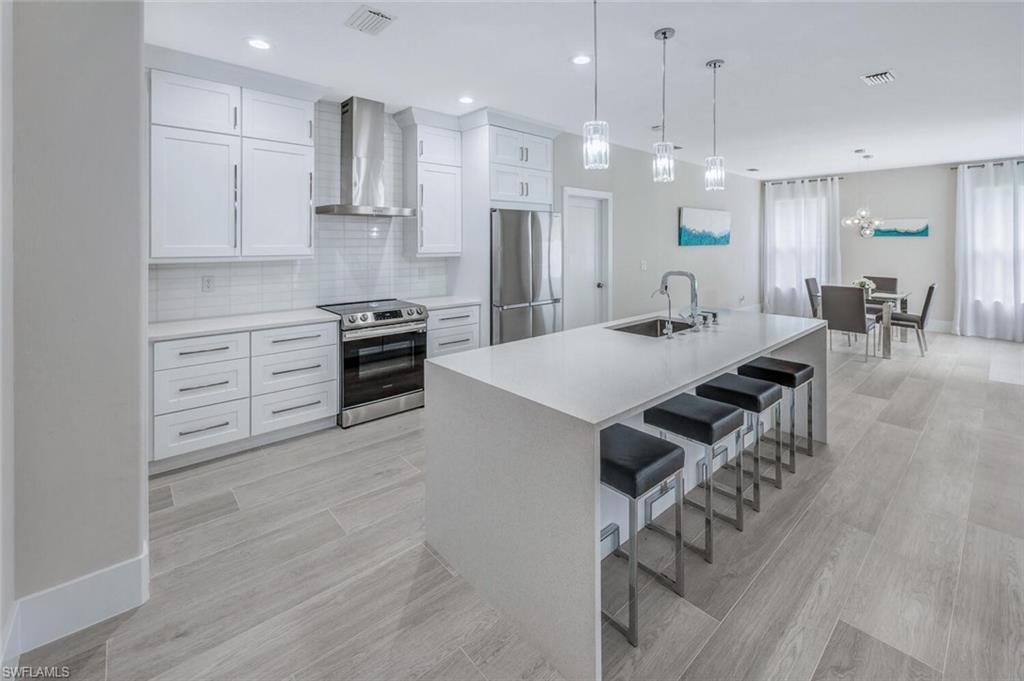 Kitchen with wall chimney exhaust hood, stainless steel appliances, white cabinets, tasteful backsplash, and decorative light fixtures