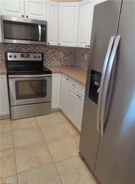 Kitchen featuring white cabinets, backsplash, stainless steel side by side door refrigerator with ice dispenser, microwave, stove & oven, backsplash, light tile patterned floors, and light countertops