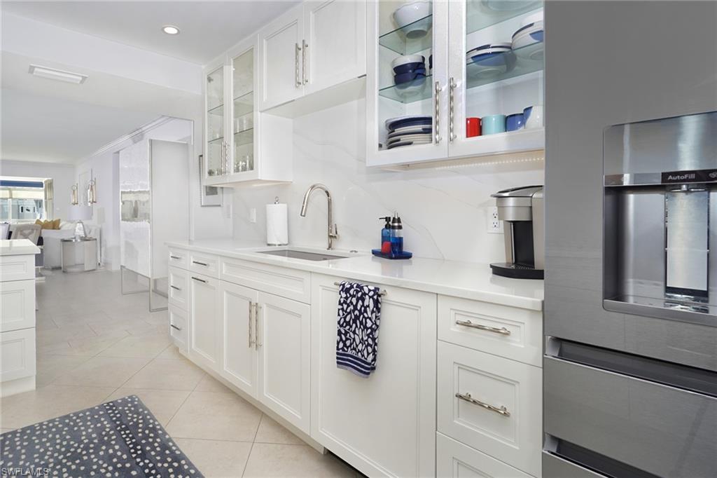 Kitchen with stainless steel fridge, glass insert cabinets, white cabinets, light tile patterned floors, and light stone counters
