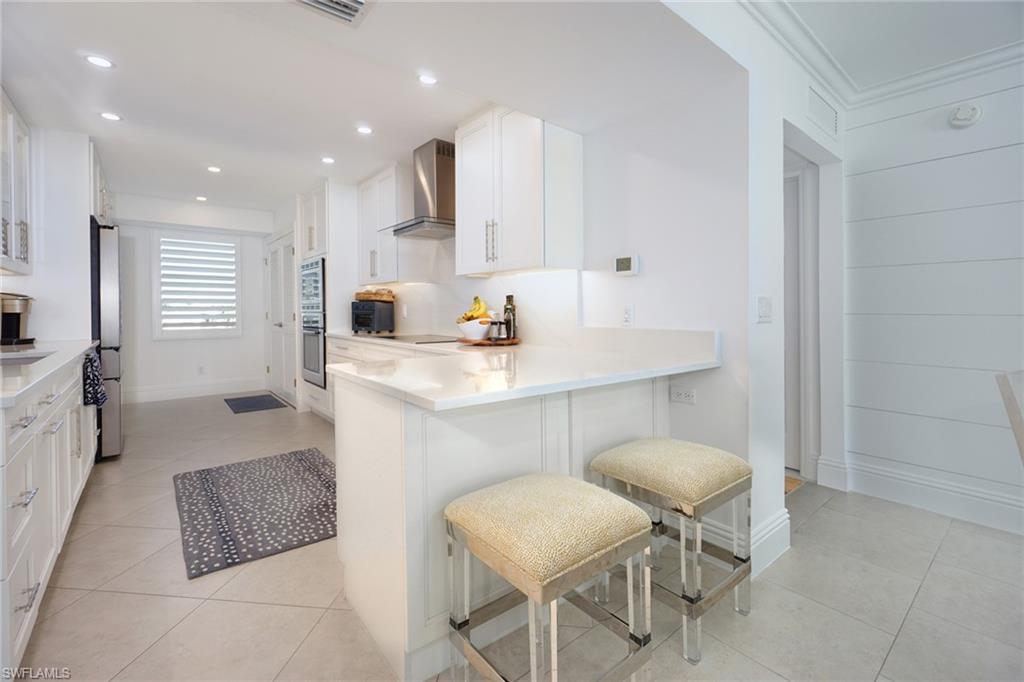 Kitchen featuring white cabinets, light stone countertops, a peninsula, wall chimney range hood, and a kitchen breakfast bar