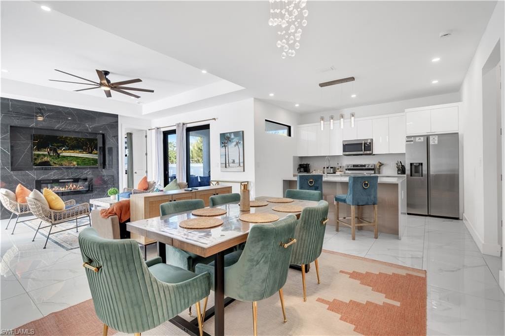 Dining room with a fireplace, light marble finish floors, recessed lighting, a tray ceiling, and a ceiling fan