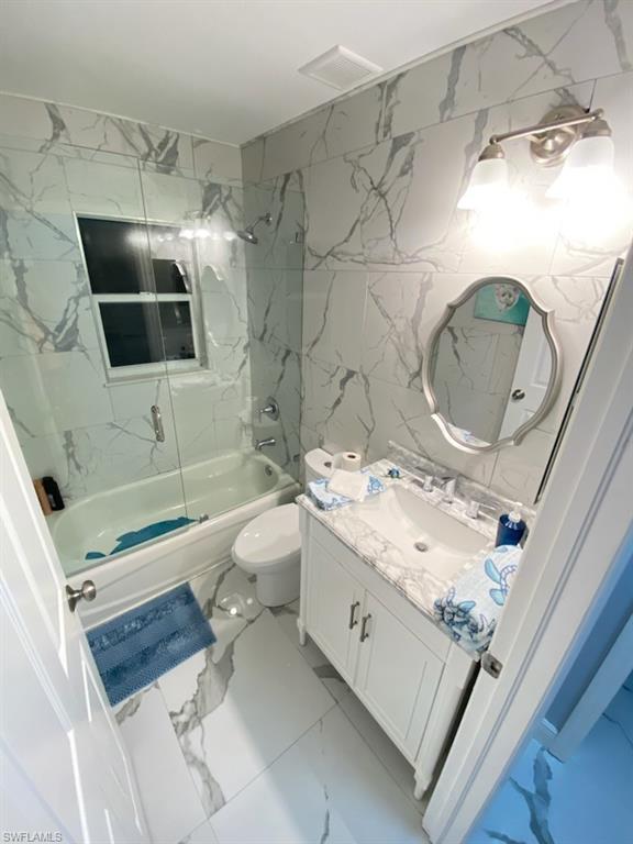 Bathroom with vanity, light marble finish floors, shower / bath combination with glass door, tile walls, and stone wall