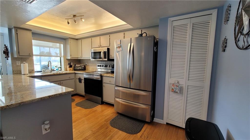 Kitchen with a sink, appliances with stainless steel finishes, light wood-type flooring, backsplash, and a tray ceiling